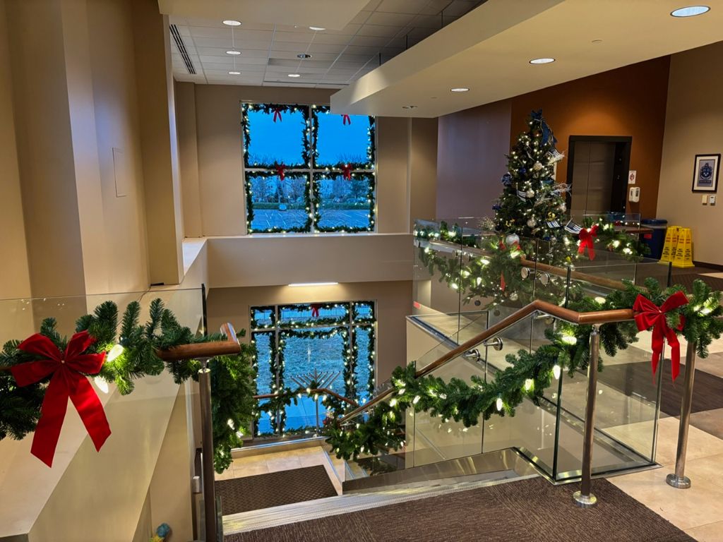 Upper landing of an open staircase with lit garland and red bows draped along the glass railings and a decorated tree nearby.