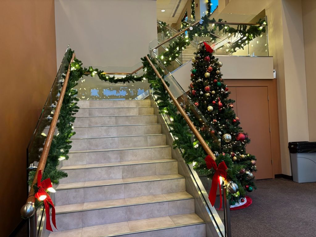 Interior staircase in a public building with lit evergreen garland along the glass railings and a decorated tree beside the stairs.