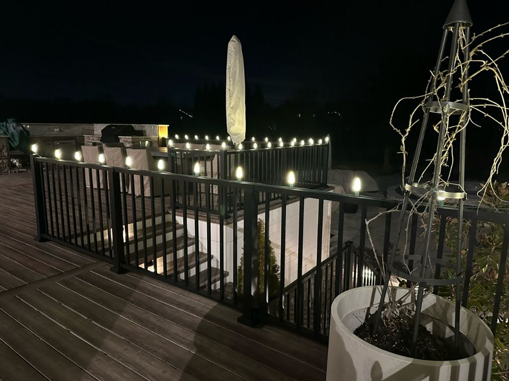Outdoor deck at night with warm white lights outlining the metal railing and a lit planter.