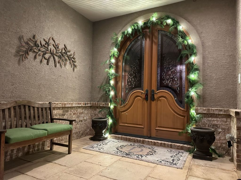 Arched double wood entry doors framed with lit evergreen garland and a bench beside the porch.