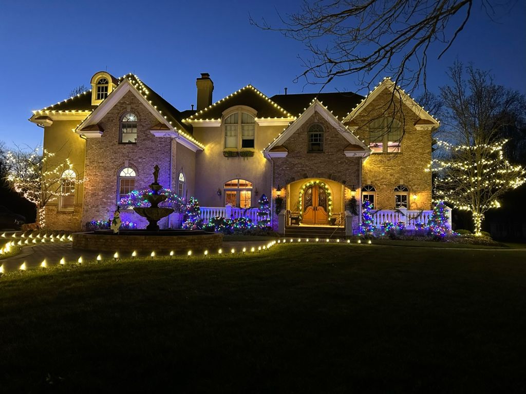 Brick and stone estate at twilight with warm white lights tracing every roofline and gable, a lit fountain and colorful blue-lit shrubs in front, wrapped trees on both sides, and staked lights lining the lawn.
