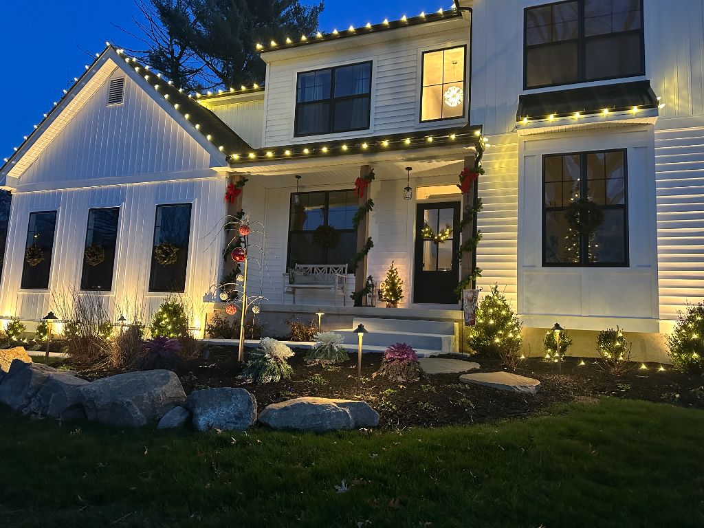Two-story white farmhouse at dusk with warm white lights along the rooflines, gables and porch, lit wreaths in the windows, and lights on the foundation shrubs and garden beds.