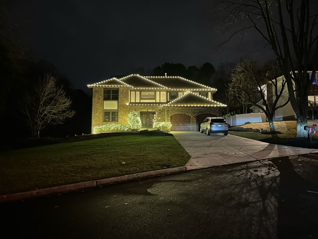 Brick two-story home at night with warm white lights tracing the rooflines and wrapped shrubs along the front of the house.