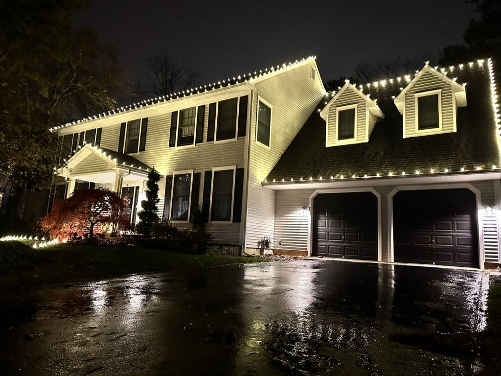 White colonial home at night with the full front roofline and dormers outlined in warm white lights above a wet driveway.