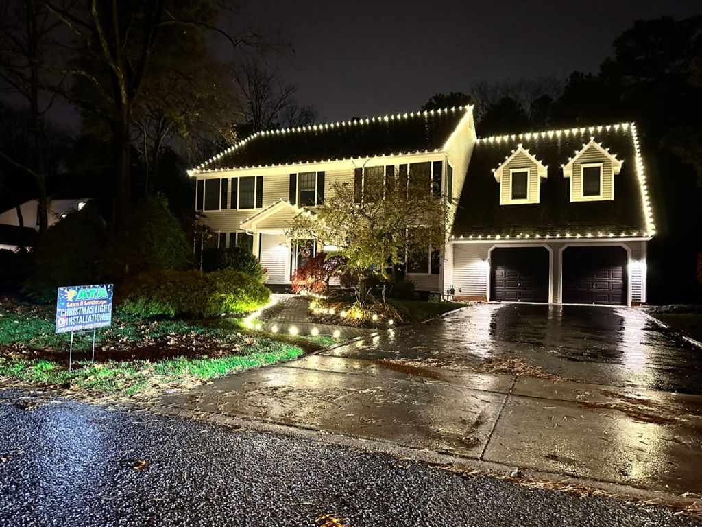 Colonial home at night with warm white roofline and dormer lights and an ATA Lawn and Landscape yard sign on the lawn.