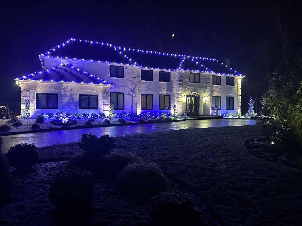 White brick home at night with blue and white lights on every roofline and blue-lit garden beds across the front.