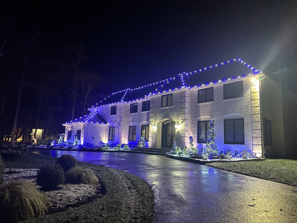 Large white brick home at night with blue and white lights outlining the roof and illuminated plantings along the drive.
