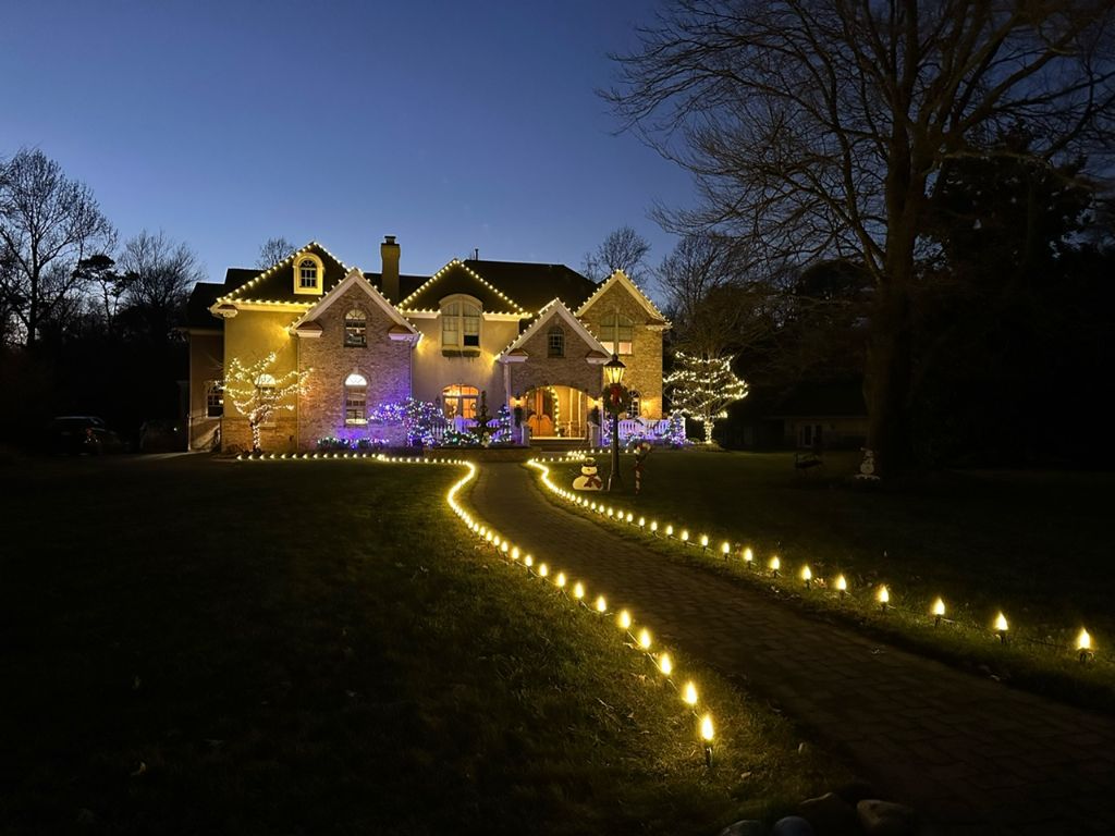 Curved driveway outlined with warm staked lights leading toward a stone home trimmed in roofline lighting at dusk.