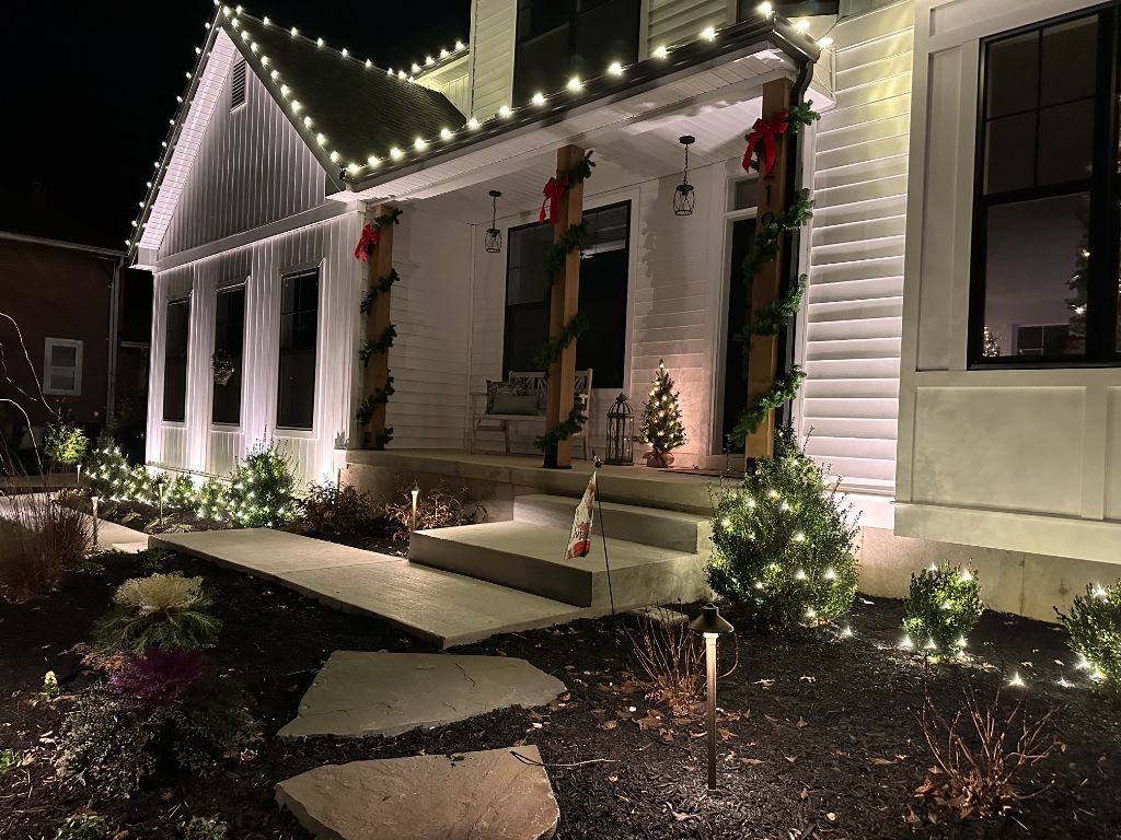 Front porch at night with warm white roofline lights, lit garland wrapped around the posts with red bows, and glowing shrubs.