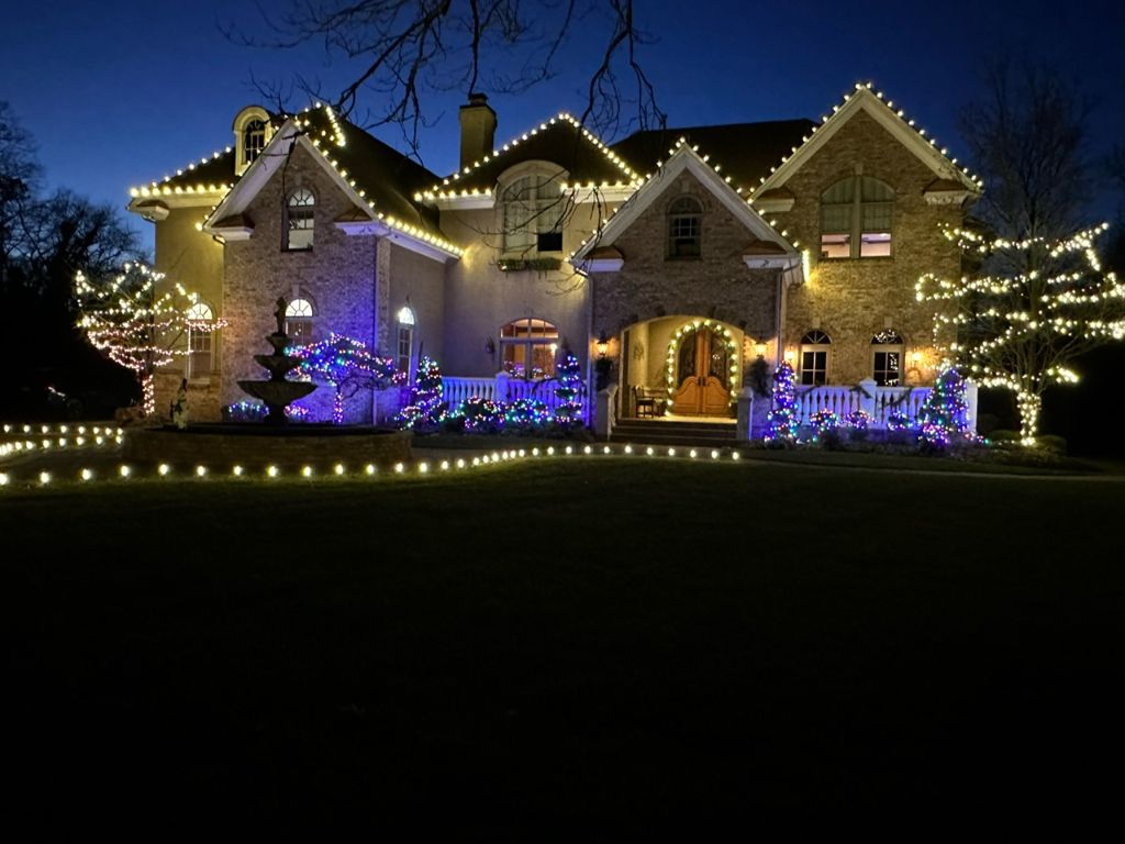 Stone estate at night with white roofline lights, blue accent lighting on shrubs and trees wrapped in lights.