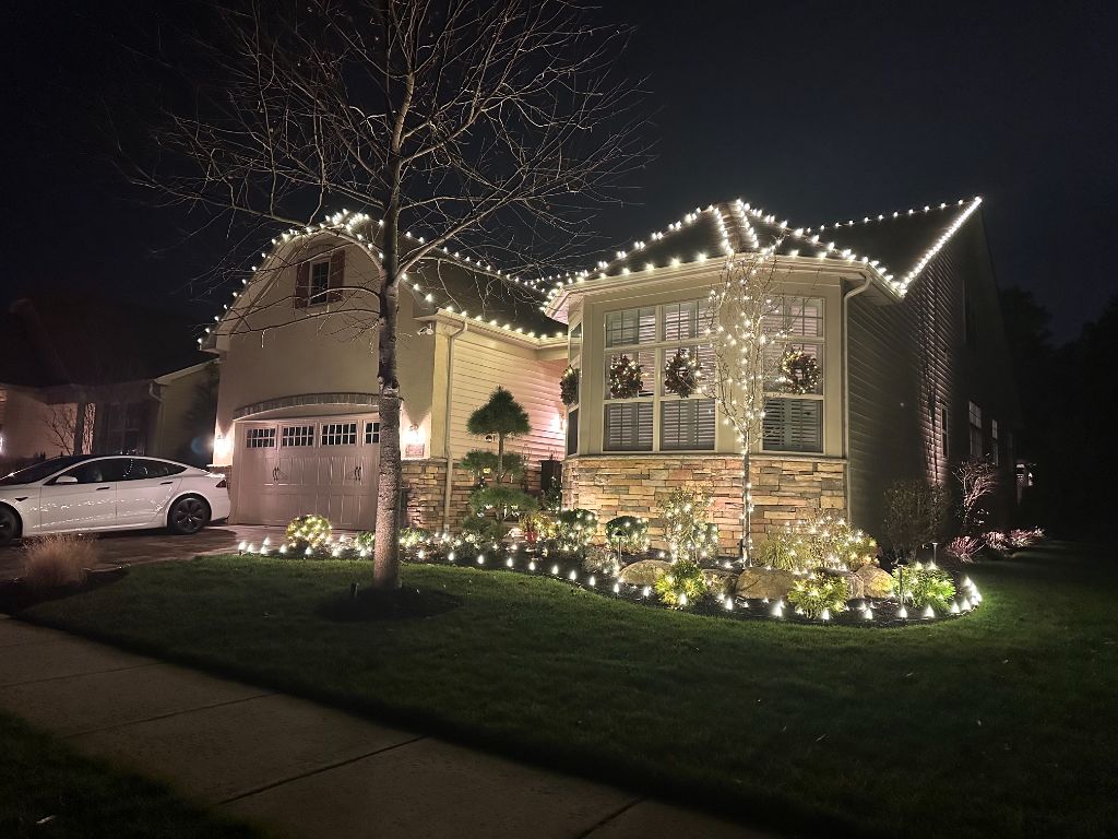 Suburban home at night with warm white lights along the gables, staked lights edging the curved garden bed, and wrapped shrubs and small trees.