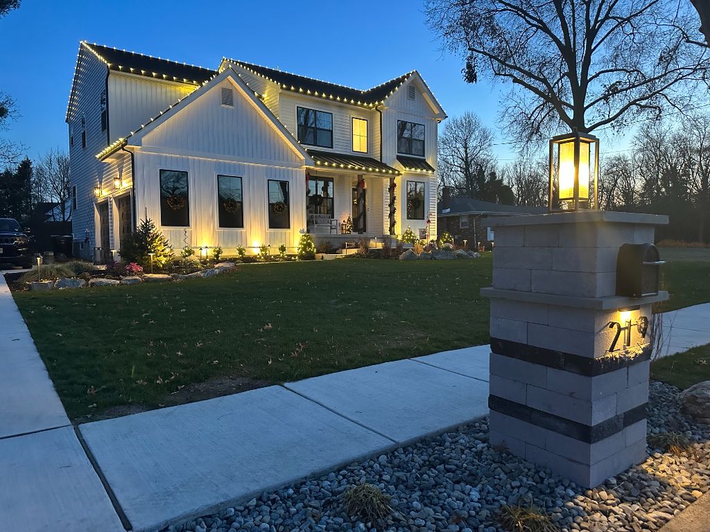 White two-story home at dusk with warm white roofline lights, lit walkway edging and a glowing lantern on a stone pillar.